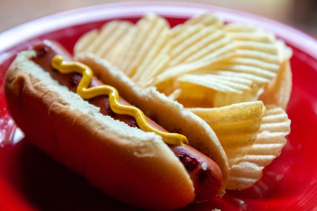 A grilled hot dog with mustard next to ridged potato chips on a red plate